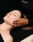 A close-up lifestyle shot of a woman receiving a facial treatment, lying down with her eyes closed while a practitioner in a black uniform wipes her face with a cotton pad. The uniform has the brand name 'Skin Accord' visible on the chest.