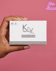 Hand holding a box of Krx Aesthetics Repair Cream against a pink background