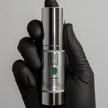 Hand wearing a black glove holding a silver and gray serum bottle labeled 'neo genesis' against a gray background.